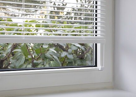 Weisse Jalousie von MHZ in Frontmontage mit Glasleistenträger am Fenster, halb geöffnet mit Blick auf Garten.