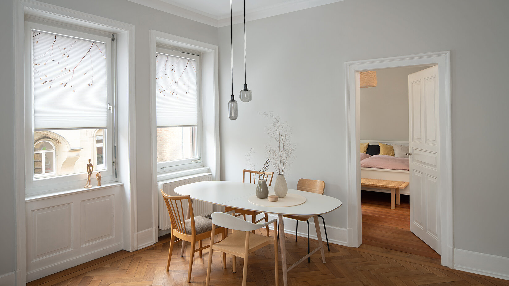 Zwei weisse Fenster mit floral bedruckten Wabenplissees in einem hellen Esszimmer mit Holzfussboden.