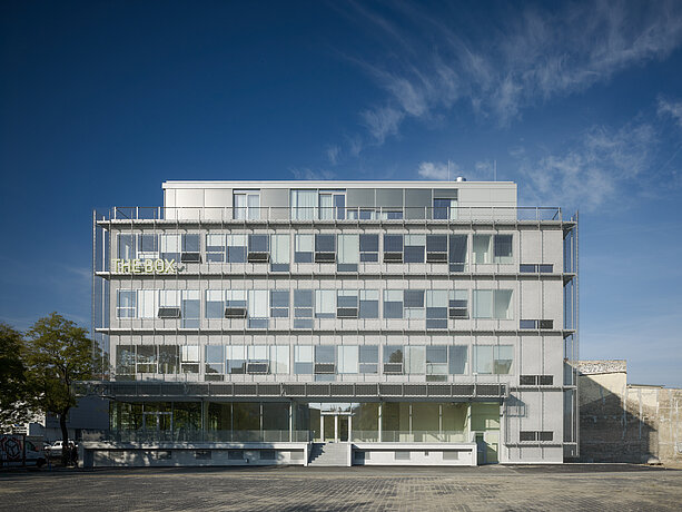 Modernes Bürogebäude mit Glasfassade und horizontalen MHZ-Sonnenschutzlamellen unter klarem Himmel.