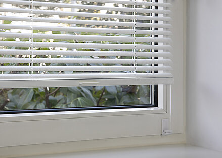MHZ TwinLine-Jalousie in Weiss mit Winkelträgermontage im Fensterfalz, halb geöffnet mit Blick ins Grüne.