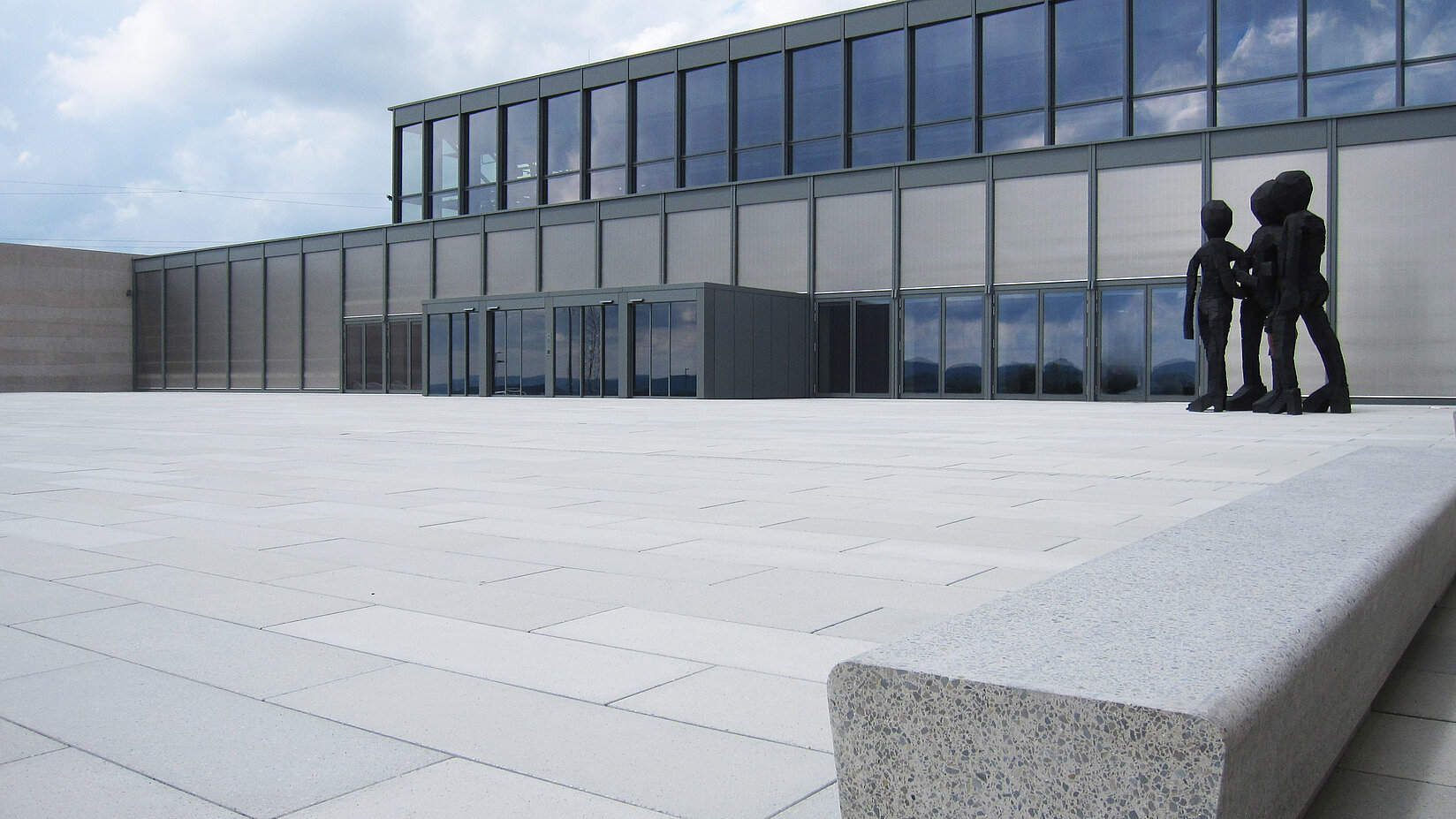Moderne Glasfassade mit grosser, leerer Vorfläche, Betonbank im Vordergrund und Menschenskulptur am rechten Bildrand.