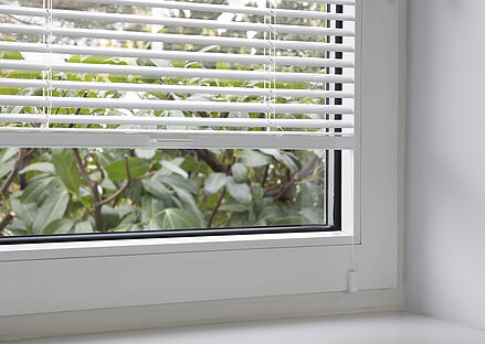Weisse Jalousie mit Fensterclipmontage am Kunststofffenster, teilweise geöffnet mit Blick ins Grüne.
