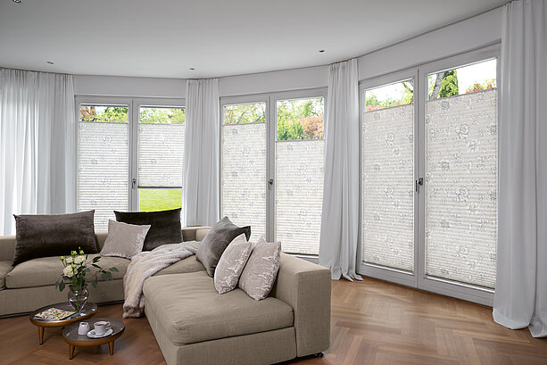 Helles Wohnzimmer mit beigem Sofa und floralen Plissees an bodentiefen Fenstern mit Blick ins Grüne.