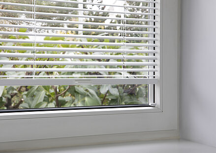 Weisse Jalousie mit MHZ-Klebeset-Montage am Fenster, halb geöffnet mit Blick ins Grüne.