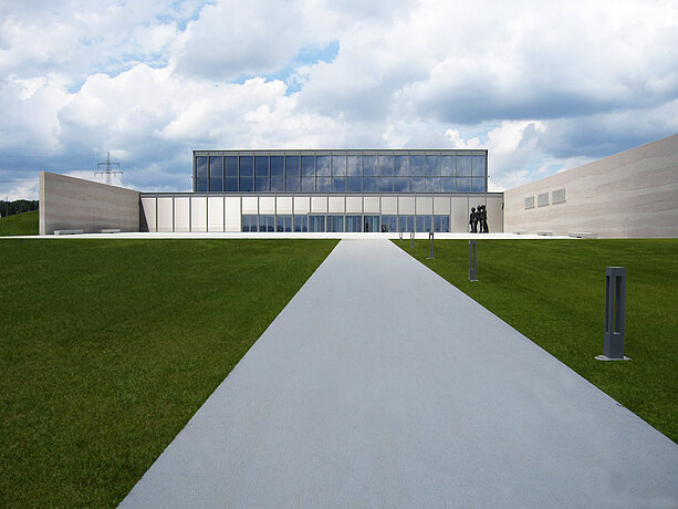 Blick entlang eines Gehwegs auf ein modernes Gebäude mit grosser Glasfassade, flankiert von Rasenflächen und mit einer Skulptur rechts vor dem Eingang.
