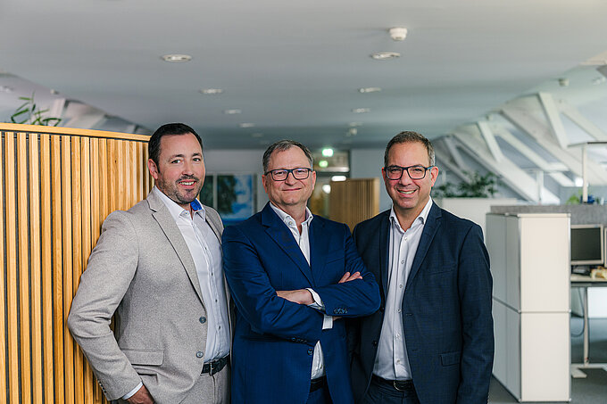 Drei Männer in Anzügen stehen in einem modernen Raum mit grossen Fenstern und Pflanzen und blicken in die Kamera.