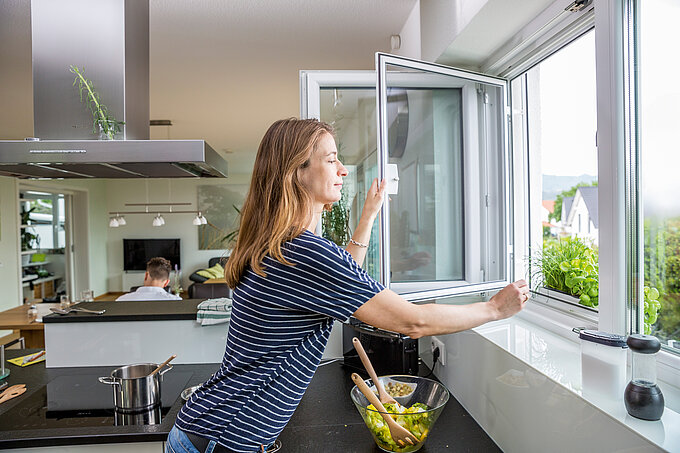 Frau öffnet ein Drehfenster mit Insektenschutz in einer modernen Küche beim Kochen.