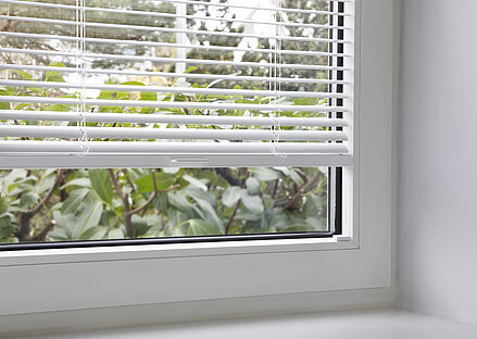 Weisse MHZ TwinLine-Jalousie mit Glasleisten-Spannschuhmontage an einem Kunststofffenster, Blick ins Grüne.