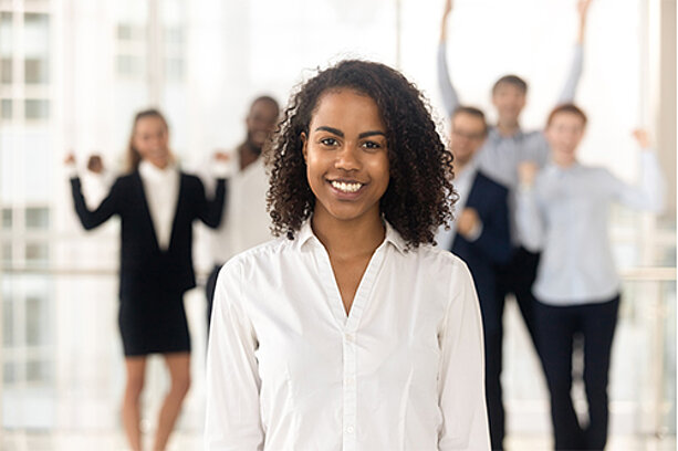 Junge Frau mit lockigem Haar steht lächelnd im Vordergrund, im Hintergrund jubelndes Team vor grossen Fensterflächen.