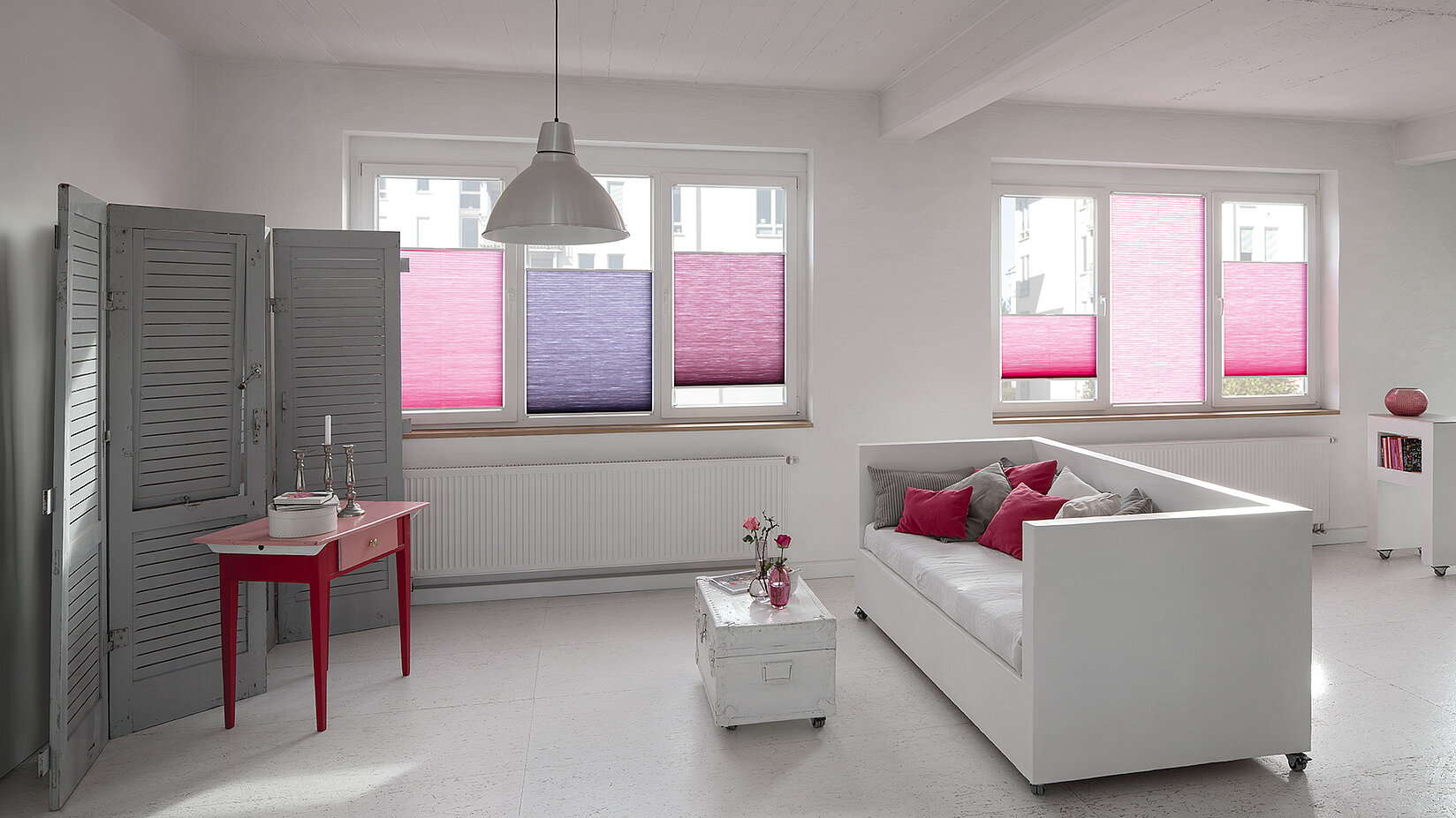 Modernes Wohnzimmer mit farbigen MHZ Duotone-Wabenplissees in Pink- und Violetttönen an weissen Fenstern.