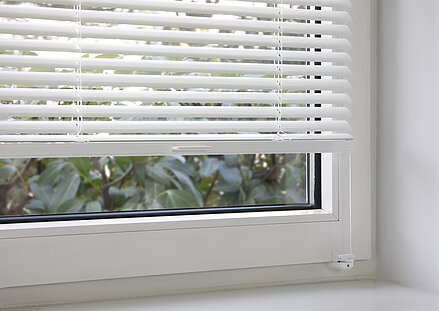 Weisse MHZ TwinLine-Jalousie mit Klemmträgermontage am Fensterflügel, halb geöffnet mit Blick nach draussen.